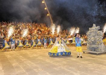 Una vivace scena festosa della Fiera Santi Popolari con artisti in costumi colorati, tra cui una donna con un abito luminoso e un uomo con un abito coordinato che regge una torcia. I fuochi d'artificio brillano intorno a loro mentre la grande folla osserva dagli spalti. Sulla destra è visibile un carro decorativo di Carvoeiro.