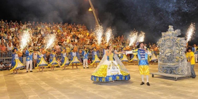 Una vivace scena festosa della Fiera Santi Popolari con artisti in costumi colorati, tra cui una donna con un abito luminoso e un uomo con un abito coordinato che regge una torcia. I fuochi d'artificio brillano intorno a loro mentre la grande folla osserva dagli spalti. Sulla destra è visibile un carro decorativo di Carvoeiro.