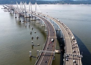 Veduta aerea del Ponte New York, un grande ponte a più corsie che attraversa un ampio fiume. Il ponte presenta un intricato design strallato e due campate separate per il traffico in direzioni opposte. Sono visibili veicoli che viaggiano su entrambi i lati, con alberi ed edifici sulla sponda opposta e una bandiera portoghese che sventola dolcemente sopra una torre.