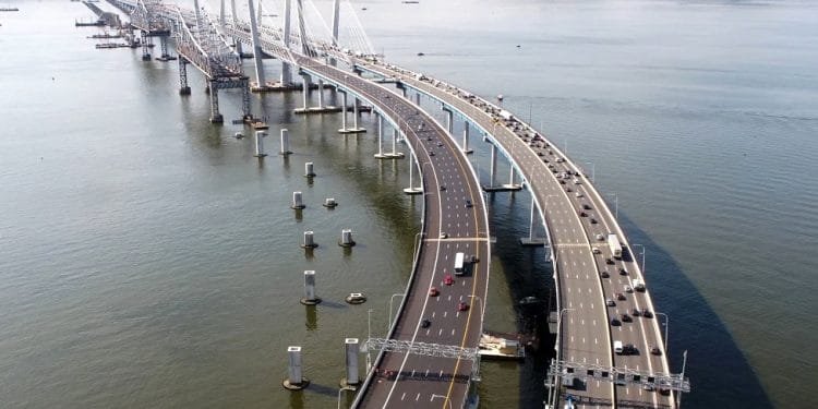 Veduta aerea del Ponte New York, un grande ponte a più corsie che attraversa un ampio fiume. Il ponte presenta un intricato design strallato e due campate separate per il traffico in direzioni opposte. Sono visibili veicoli che viaggiano su entrambi i lati, con alberi ed edifici sulla sponda opposta e una bandiera portoghese che sventola dolcemente sopra una torre.