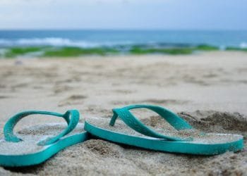 Un paio di infradito verde acqua giacciono semisepolte nella sabbia su una spiaggia in Portogallo, con l'oceano blu e un cielo nuvoloso sullo sfondo. Rade alghe verdi possono essere viste sulla spiaggia sabbiosa vicino all'acqua.