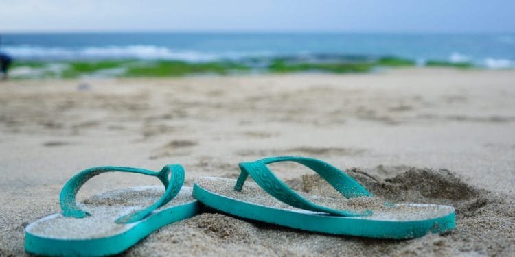 Un paio di infradito verde acqua giacciono semisepolte nella sabbia su una spiaggia in Portogallo, con l'oceano blu e un cielo nuvoloso sullo sfondo. Rade alghe verdi possono essere viste sulla spiaggia sabbiosa vicino all'acqua.