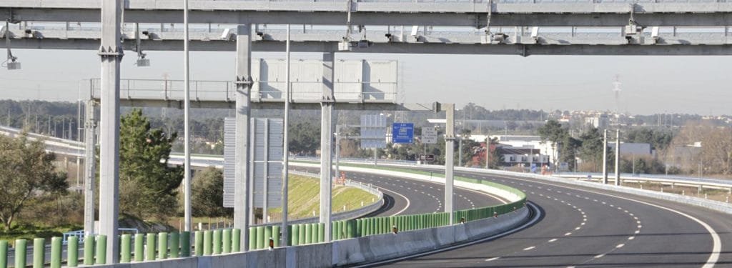 Una chiara autostrada a più corsie curva dolcemente a sinistra, priva di veicoli. In alto ci sono diverse strutture metalliche con cartelli che indicano i limiti del portogallo e le tariffe dei pedaggi. Il paesaggio comprende alcuni edifici, alberi e un paesaggio urbano distante sotto un cielo limpido.