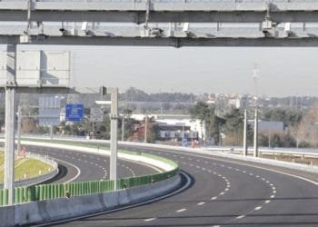 Una chiara autostrada a più corsie curva dolcemente a sinistra, priva di veicoli. In alto ci sono diverse strutture metalliche con cartelli che indicano i limiti del portogallo e le tariffe dei pedaggi. Il paesaggio comprende alcuni edifici, alberi e un paesaggio urbano distante sotto un cielo limpido.