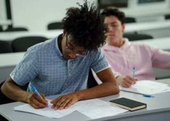 Due studenti sono seduti in un’aula. Lo studente in primo piano, che indossa una camicia a quadri blu, si concentra sulla scrittura su un foglio. Lo studente sullo sfondo, vestito con una felpa con cappuccio rosa, osserva lo studente in primo piano. Entrambi hanno penne e fogli sulle loro scrivanie, che mostrano scene tipiche delle scuole pubbliche classificate nazionali.