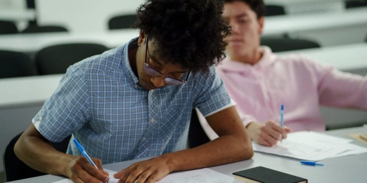 Due studenti sono seduti in un’aula. Lo studente in primo piano, che indossa una camicia a quadri blu, si concentra sulla scrittura su un foglio. Lo studente sullo sfondo, vestito con una felpa con cappuccio rosa, osserva lo studente in primo piano. Entrambi hanno penne e fogli sulle loro scrivanie, che mostrano scene tipiche delle scuole pubbliche classificate nazionali.