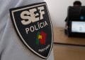 Primo piano di un distintivo del Servizio Stranieri Frontiere SEF Polícia su un'uniforme, con la bandiera del Portogallo inclusa sul distintivo. Sullo sfondo sfocato, una persona è seduta alla scrivania con un laptop dallo schermo indistinguibile.