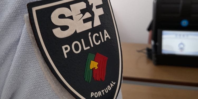 Primo piano di un distintivo del Servizio Stranieri Frontiere SEF Polícia su un'uniforme, con la bandiera del Portogallo inclusa sul distintivo. Sullo sfondo sfocato, una persona è seduta alla scrivania con un laptop dallo schermo indistinguibile.