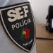 Primo piano di un distintivo del Servizio Stranieri Frontiere SEF Polícia su un'uniforme, con la bandiera del Portogallo inclusa sul distintivo. Sullo sfondo sfocato, una persona è seduta alla scrivania con un laptop dallo schermo indistinguibile.