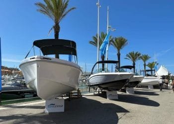 Una schiera di barche è in mostra a Marina Vilamoura sotto un cielo azzurro e limpido. Sullo sfondo sono visibili palme e barche ormeggiate. Davanti a ogni barca sono posizionati cartelli con la scritta "BOAT CENTER" mentre i visitatori si godono il International Boat Show camminando e osservando le imponenti navi.