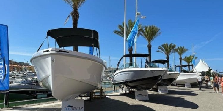 Una schiera di barche è in mostra a Marina Vilamoura sotto un cielo azzurro e limpido. Sullo sfondo sono visibili palme e barche ormeggiate. Davanti a ogni barca sono posizionati cartelli con la scritta "BOAT CENTER" mentre i visitatori si godono il International Boat Show camminando e osservando le imponenti navi.