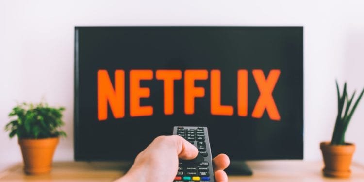 Una mano che regge un telecomando è puntata verso un televisore che mostra il logo Netflix, leader dello streaming in Portogallo. La TV è su un supporto di legno con piante in vaso su entrambi i lati.