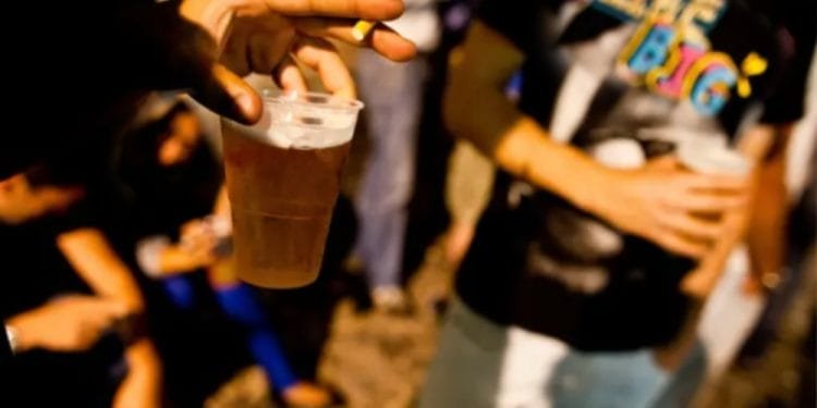 Una persona che tiene una sigaretta e una tazza di birra in una mano, con un gruppo di individui sfocati sullo sfondo. La persona in primo piano indossa una maglietta nera con un testo colorato che dice "I LIKE BIG". La scena sembra essere un incontro sociale, forse reminiscenza della vita notturna in Portogallo.