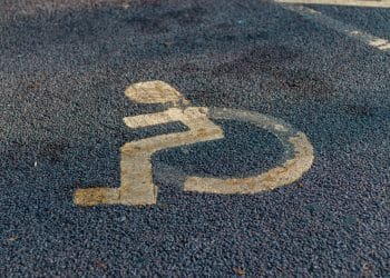 Su una superficie asfaltata è dipinto il simbolo sbiadito di una sedia a rotelle, che indica uno spazio di parcheggio accessibile designato per le persone con disabilità. Lo sfondo è grigio e strutturato, che ricorda applicazioni simili di segnaletica di parcheggio viste in luoghi come il Portogallo.