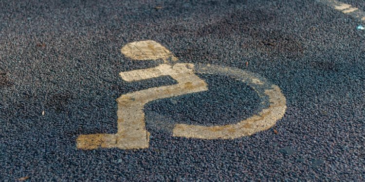 Su una superficie asfaltata è dipinto il simbolo sbiadito di una sedia a rotelle, che indica uno spazio di parcheggio accessibile designato per le persone con disabilità. Lo sfondo è grigio e strutturato, che ricorda applicazioni simili di segnaletica di parcheggio viste in luoghi come il Portogallo.