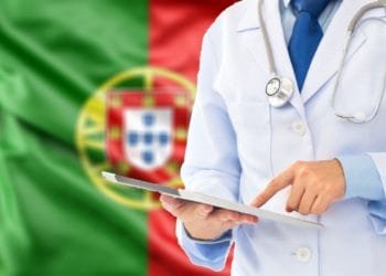 Una persona che indossa un camice bianco e uno stetoscopio usa un tablet. Sullo sfondo è riprodotta la bandiera del Portogallo, con campi verdi e rossi e lo stemma nazionale, che evidenzia l'assistenza sanitaria portogallo.