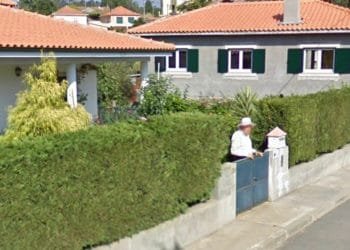 Una persona si trova davanti al cancello di una casa di cemento con un tetto di tegole rosse, delimitata da una siepe verde lussureggiante. Sullo sfondo è visibile un'altra casa con copertura simile. Questa scena serena, che ricorda una cattura di Google Maps, suggerisce un tranquillo quartiere residenziale in una giornata soleggiata.