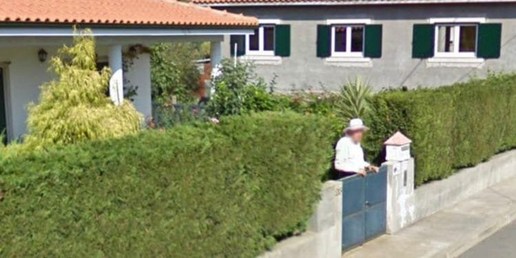 Una persona si trova davanti al cancello di una casa di cemento con un tetto di tegole rosse, delimitata da una siepe verde lussureggiante. Sullo sfondo è visibile un'altra casa con copertura simile. Questa scena serena, che ricorda una cattura di Google Maps, suggerisce un tranquillo quartiere residenziale in una giornata soleggiata.
