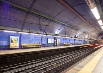 Una spaziosa piattaforma sotterranea della metropolitana con piastrelle a mosaico blu sulle pareti e sul soffitto del sistema metropolitano di Lisbona. Il posto è in gran parte vuoto, fatta eccezione per poche persone in lontananza. I binari attraversano il centro e sulla banchina ci sono panchine e manifesti pubblicitari.