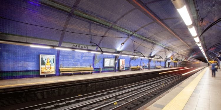 Una spaziosa piattaforma sotterranea della metropolitana con piastrelle a mosaico blu sulle pareti e sul soffitto del sistema metropolitano di Lisbona. Il posto è in gran parte vuoto, fatta eccezione per poche persone in lontananza. I binari attraversano il centro e sulla banchina ci sono panchine e manifesti pubblicitari.