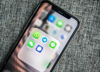 Lo schermo di uno smartphone che mostra una cartella denominata "Messaggi" contenente app di messaggistica come WhatsApp, Telegram, LINE, WeChat e Signal poggia su una superficie grigia e strutturata. Tra le icone delle app in verde, blu e grigio con vari design c'è il mini-programma WeChat di Turismo Portugal lancia.