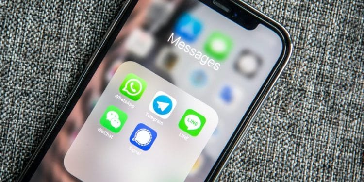 Lo schermo di uno smartphone che mostra una cartella denominata "Messaggi" contenente app di messaggistica come WhatsApp, Telegram, LINE, WeChat e Signal poggia su una superficie grigia e strutturata. Tra le icone delle app in verde, blu e grigio con vari design c'è il mini-programma WeChat di Turismo Portugal lancia.