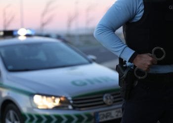 Un agente delle forze dell'ordine in uniforme tiene le manette, in piedi accanto a un'auto di pattuglia con luci lampeggianti. L'auto, contrassegnata con la scritta "GNR" sul cofano, è parcheggiata sul ciglio della strada durante il crepuscolo. Lo sfondo presenta una vista sfocata di alberi e un cielo pallido, alludendo alla loro vigilanza sui trasgressori delle disposizioni doganali.