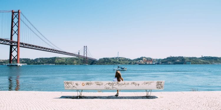 Una persona si siede su una panchina ricoperta di graffiti di fronte a un grande ponte sospeso rosso che attraversa uno specchio d'acqua calmo, con colline verdi ed edifici sullo sfondo sotto un cielo azzurro, forse riflettendo sulle frequenti domande di trasferimento in Portogallo.