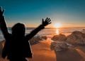 Una persona con le braccia alzate si staglia contro un vibrante tramonto su una spiaggia, sognando di trasferirsi in Portogallo. Il sole è basso sull'orizzonte, proiettando una calda luce sulla sabbia e sulle rocce sparse lungo la costa. Il cielo è una sfumatura di colori dal blu intenso all'arancione.