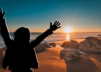 Una persona con le braccia alzate si staglia contro un vibrante tramonto su una spiaggia, sognando di trasferirsi in Portogallo. Il sole è basso sull'orizzonte, proiettando una calda luce sulla sabbia e sulle rocce sparse lungo la costa. Il cielo è una sfumatura di colori dal blu intenso all'arancione.