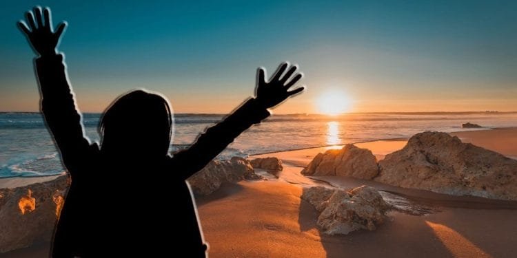 Una persona con le braccia alzate si staglia contro un vibrante tramonto su una spiaggia, sognando di trasferirsi in Portogallo. Il sole è basso sull'orizzonte, proiettando una calda luce sulla sabbia e sulle rocce sparse lungo la costa. Il cielo è una sfumatura di colori dal blu intenso all'arancione.