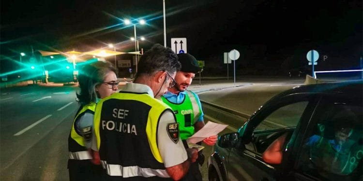 Tre agenti di polizia, indossando giubbotti ad alta visibilità, effettuano un controllo stradale notturno. Due agenti esaminano i documenti mentre un altro resta a guardare. Si vede un conducente di un'auto nera interagire con gli agenti durante i controlli frontiere portoghesi per la GMG. La scena è illuminata dai lampioni.