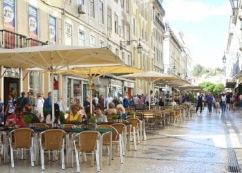 Una vivace scena del Costo della vita in Portogallo. Questa strada a Lisbona con persone sedute ai tavolini di un bar all'aperto sotto grandi ombrelloni gialli. L'ampia strada pedonale, è fiancheggiata da edifici storici con negozi e balconi.