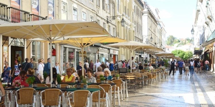 Una vivace scena del Costo della vita in Portogallo. Questa strada a Lisbona con persone sedute ai tavolini di un bar all'aperto sotto grandi ombrelloni gialli. L'ampia strada pedonale, è fiancheggiata da edifici storici con negozi e balconi.