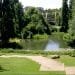 Una vista pittoresca di un parco caratterizzato da un tranquillo laghetto circondato da una vegetazione lussureggiante. Gli alberi incorniciano la scena, con una scalinata che scende alla zona erbosa vicino al laghetto. Si vede una persona pescare in riva all'acqua. Gli edifici fanno capolino tra gli alberi sullo sfondo, ricordando il fascino dei Giardini Gulbenkian.