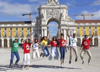 Un gruppo di sette giovani salta gioiosamente in aria, sorridendo e tenendosi per mano. Si trovano in una grande piazza con un arco storico ed edifici colorati sullo sfondo. Alcuni indossano magliette colorate con il logo di un cuore, celebrando la Giornata Mondiale Gioventù con gli amici italiani.