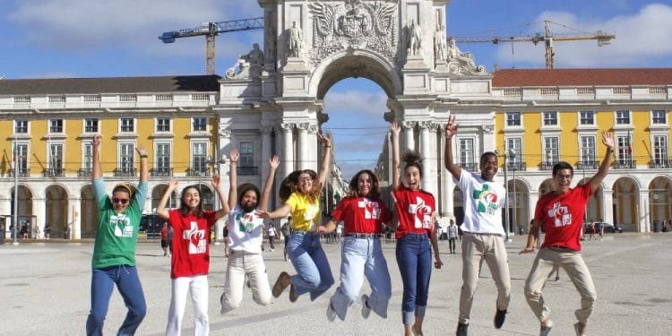 Un gruppo di sette giovani salta gioiosamente in aria, sorridendo e tenendosi per mano. Si trovano in una grande piazza con un arco storico ed edifici colorati sullo sfondo. Alcuni indossano magliette colorate con il logo di un cuore, celebrando la Giornata Mondiale Gioventù con gli amici italiani.
