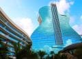Un edificio moderno progettato per assomigliare ad una chitarra blu brillante si staglia contro un cielo parzialmente nuvoloso. Con finestre in vetro riflettente, l'Hard Rock Hotel Algarve trasuda eleganza contemporanea. Un altro edificio curvo e le palme esaltano l'atmosfera tropicale in primo piano.