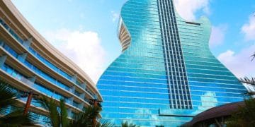 Un edificio moderno progettato per assomigliare ad una chitarra blu brillante si staglia contro un cielo parzialmente nuvoloso. Con finestre in vetro riflettente, l'Hard Rock Hotel Algarve trasuda eleganza contemporanea. Un altro edificio curvo e le palme esaltano l'atmosfera tropicale in primo piano.