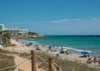 Una vivace scena sulla spiaggia con persone che prendono il sole, nuotano e si rilassano sotto ombrelloni colorati. Il mare cristallino incontra l'arenile sabbioso, mentre l'Hotel Adulti si affaccia sulla spiaggia da sinistra, sullo sfondo di un cielo terso e soleggiato.