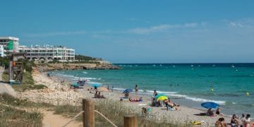 Una vivace scena sulla spiaggia con persone che prendono il sole, nuotano e si rilassano sotto ombrelloni colorati. Il mare cristallino incontra l'arenile sabbioso, mentre l'Hotel Adulti si affaccia sulla spiaggia da sinistra, sullo sfondo di un cielo terso e soleggiato.