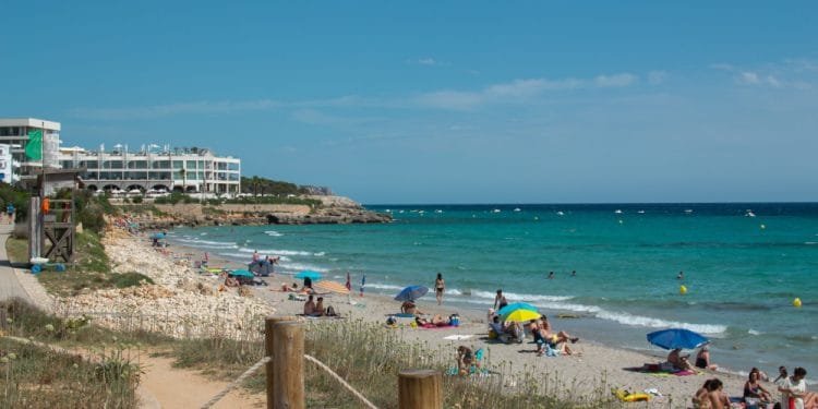 Una vivace scena sulla spiaggia con persone che prendono il sole, nuotano e si rilassano sotto ombrelloni colorati. Il mare cristallino incontra l'arenile sabbioso, mentre l'Hotel Adulti si affaccia sulla spiaggia da sinistra, sullo sfondo di un cielo terso e soleggiato.