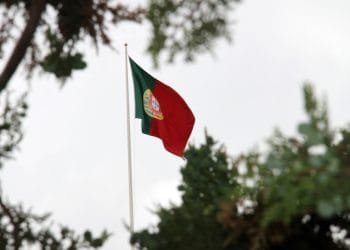 Una bandiera portoghese con sezioni verdi e rosse e uno stemma al centro sventola su un pennone contro un cielo nuvoloso, circondata da rami di alberi in Portogallo, che incarna il grado della nazione nel Global Peace Index.