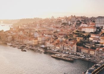 Veduta aerea di una pittoresca città lungo il fiume al tramonto. Il paesaggio urbano presenta un mix di edifici storici con tetti in terracotta, strade tortuose e un fiume fiancheggiato da barche e moli. Attraverso Strategie Abitative Locali, la tenue tonalità dorata del cielo aggiunge calore alla scena pittoresca.