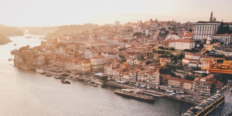 Veduta aerea di una pittoresca città lungo il fiume al tramonto. Il paesaggio urbano presenta un mix di edifici storici con tetti in terracotta, strade tortuose e un fiume fiancheggiato da barche e moli. Attraverso Strategie Abitative Locali, la tenue tonalità dorata del cielo aggiunge calore alla scena pittoresca.