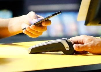 Una persona utilizza uno smartphone per effettuare un pagamento contactless presso un terminale da banco, mentre un'altra persona tiene fermo il terminale di pagamento, probabilmente garantendo che il processo proceda senza intoppi. Entrambe le mani sono mostrate su una superficie di colore giallo, evitando ogni possibilità di rimanere intrappolati in una truffa di Rete Criminale Truffe MbWay.