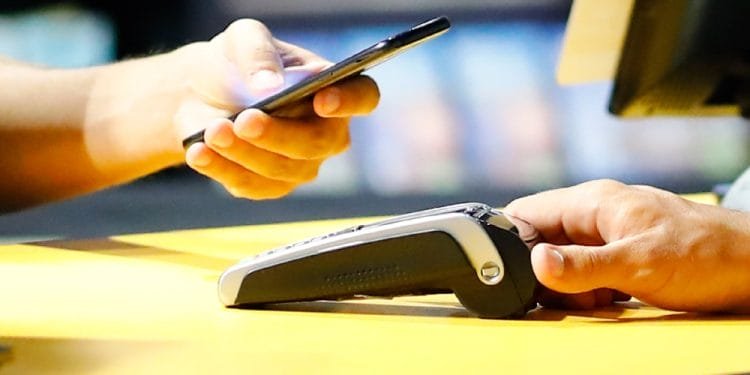 Una persona utilizza uno smartphone per effettuare un pagamento contactless presso un terminale da banco, mentre un'altra persona tiene fermo il terminale di pagamento, probabilmente garantendo che il processo proceda senza intoppi. Entrambe le mani sono mostrate su una superficie di colore giallo, evitando ogni possibilità di rimanere intrappolati in una truffa di Rete Criminale Truffe MbWay.
