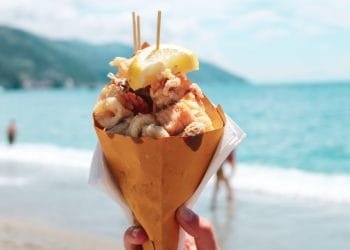 Una mano tiene un cono di carta ripieno di frutti di mare fritti e sormontato da uno spicchio di limone sullo sfondo di una spiaggia con acqua blu e montagne lontane. Sullo sfondo si vedono alcune persone che si godono la spiaggia, magari sognando di ordinare delizie simili da consegnare tramite Uber Eats.