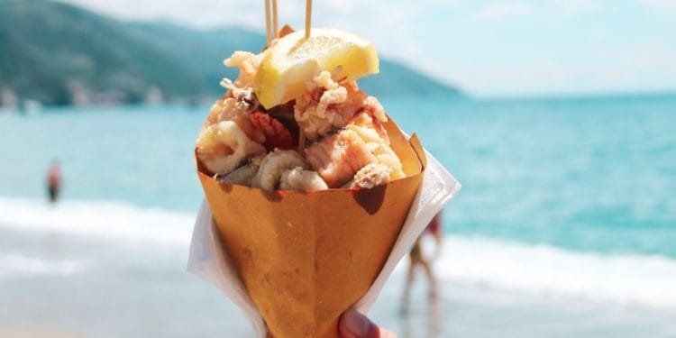 Una mano tiene un cono di carta ripieno di frutti di mare fritti e sormontato da uno spicchio di limone sullo sfondo di una spiaggia con acqua blu e montagne lontane. Sullo sfondo si vedono alcune persone che si godono la spiaggia, magari sognando di ordinare delizie simili da consegnare tramite Uber Eats.