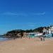 Una spiaggia sabbiosa con poche persone che camminano lungo la riva e nell'acqua. Edifici colorati e ombrelloni sono visibili sulla destra, vicino alle scale che conducono alla spiaggia. Il cielo è limpido e azzurro, con alcuni alberi e scogliere sullo sfondo, che ricordano le tranquille spiagge dell'Algarve dove l'accesso a volte può essere vietato.